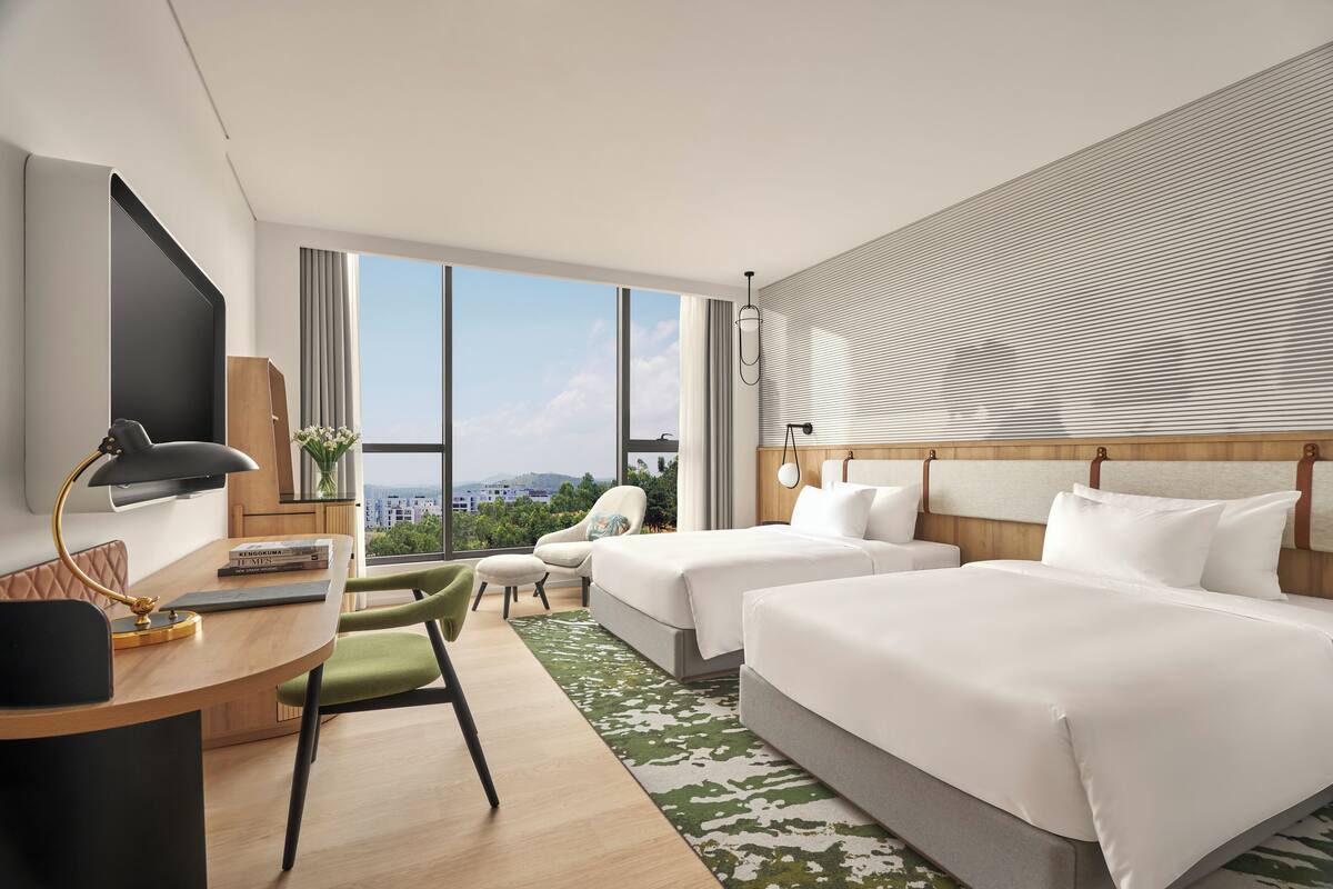 Habitación de hotel moderna con dos camas, un escritorio, una silla y un amplio ventanal con vista a la ciudad.