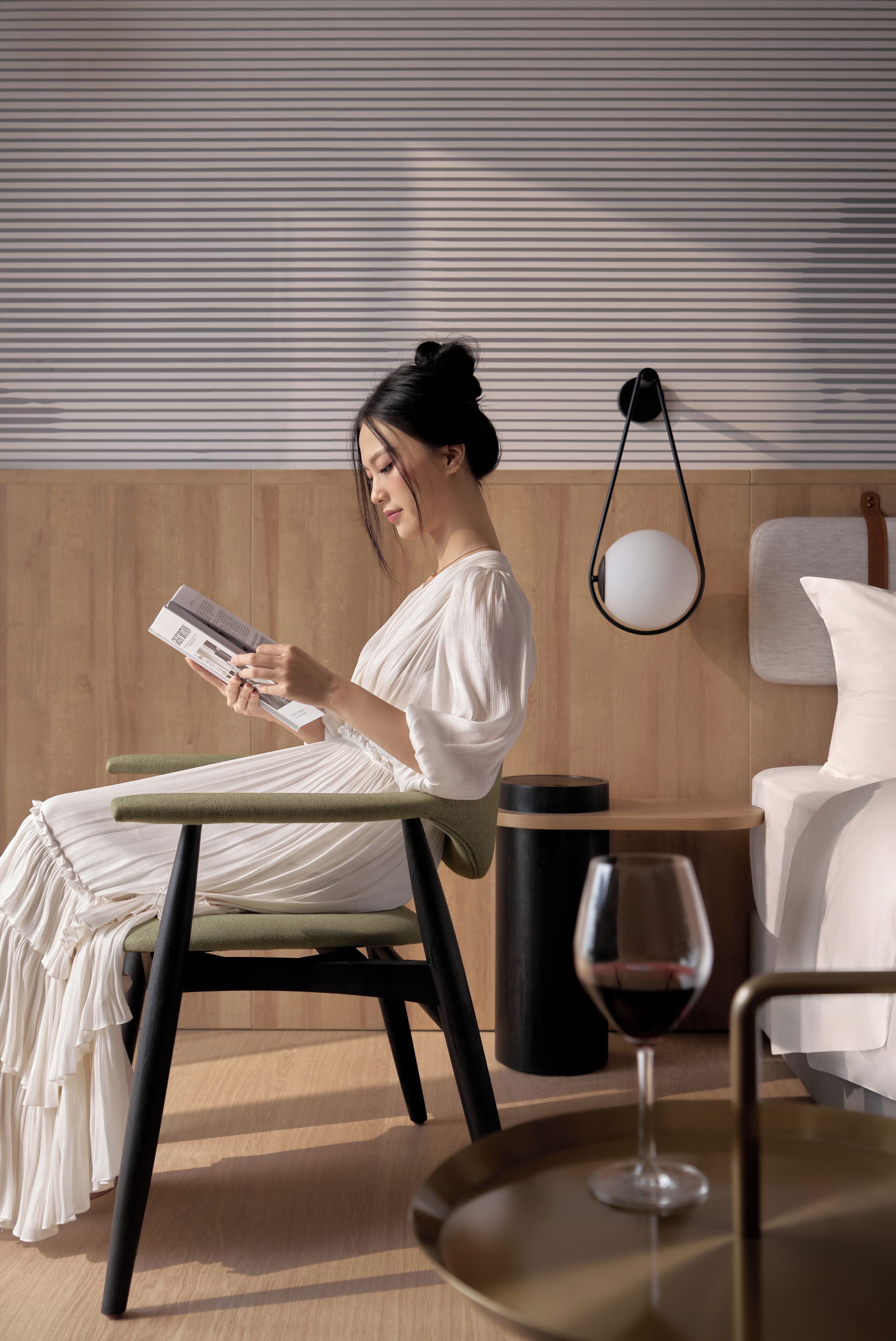 A person reading a book in a modern room with a wooden panel wall, a bedside table with a lamp, and a glass of red wine.