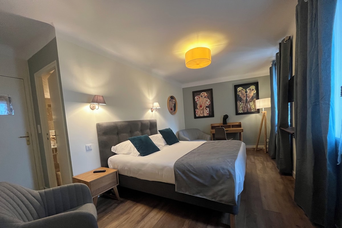 A hotel room with a large bed, a bedside table, a chair, and a desk with a lamp.