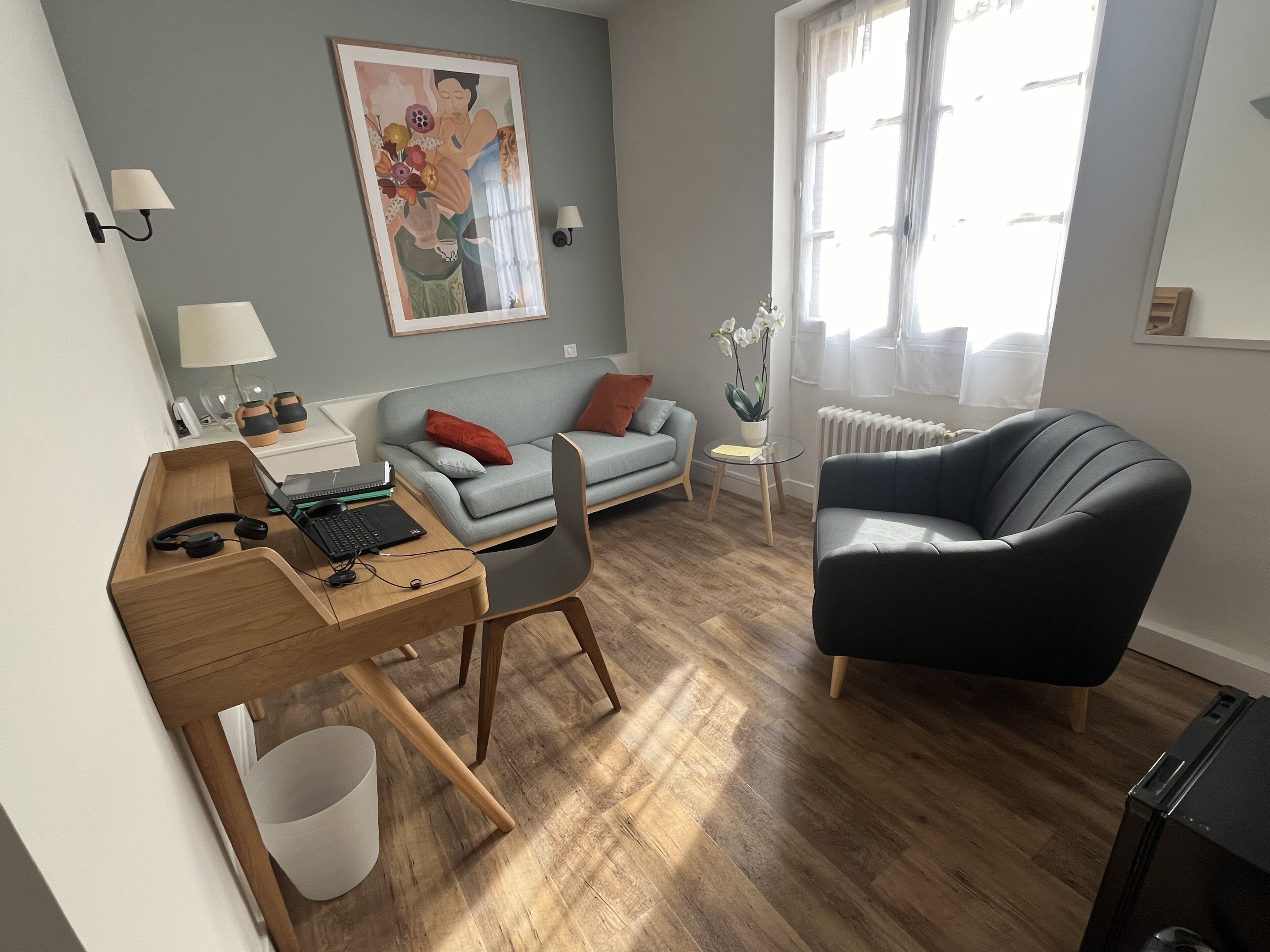 A modern living room with a wooden desk, a grey sofa, a black armchair, and a painting on the wall.