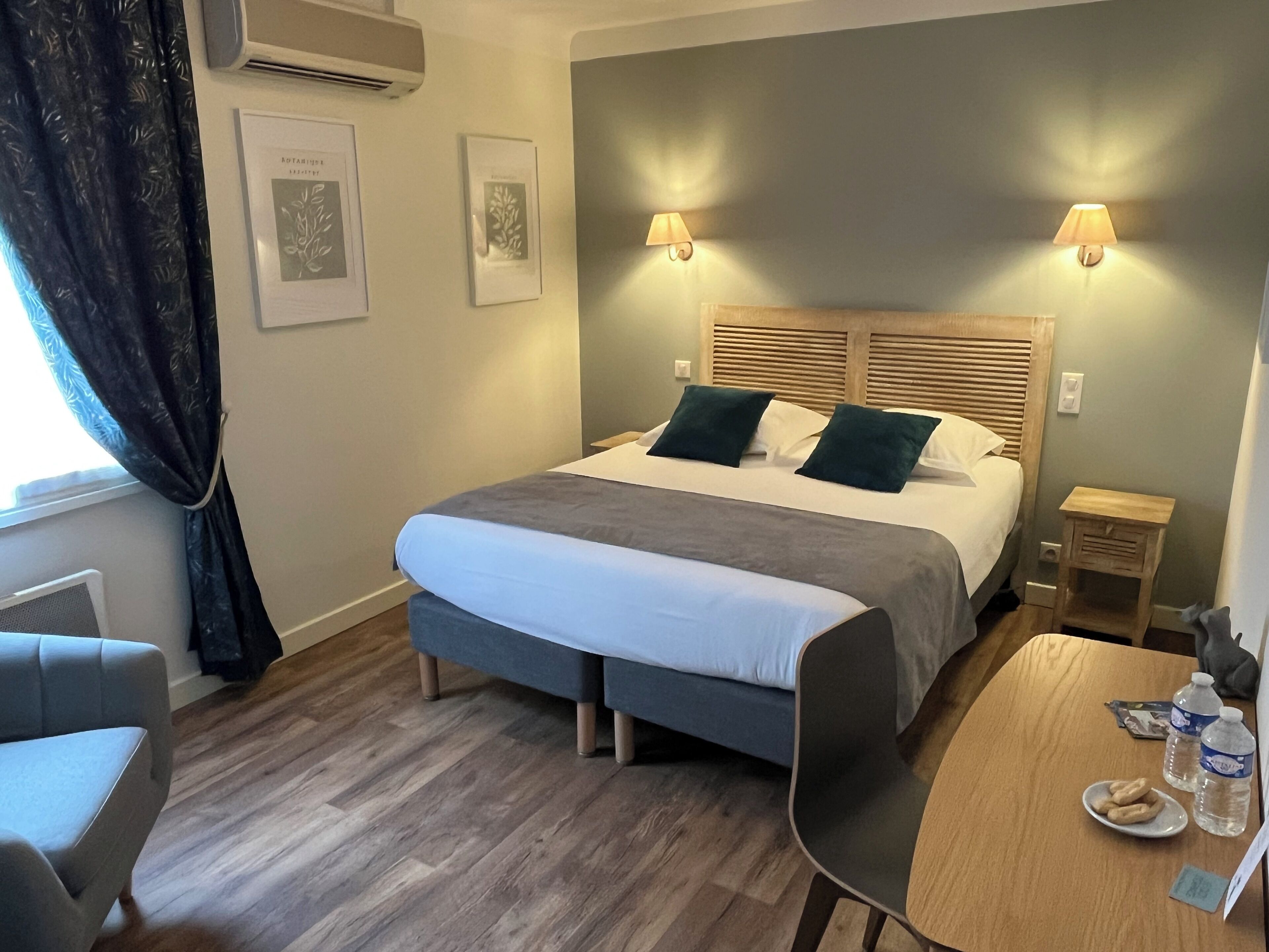A modern hotel room with a large bed, two bedside lamps, a wooden headboard, a small table with a plate of cookies, and a chair.