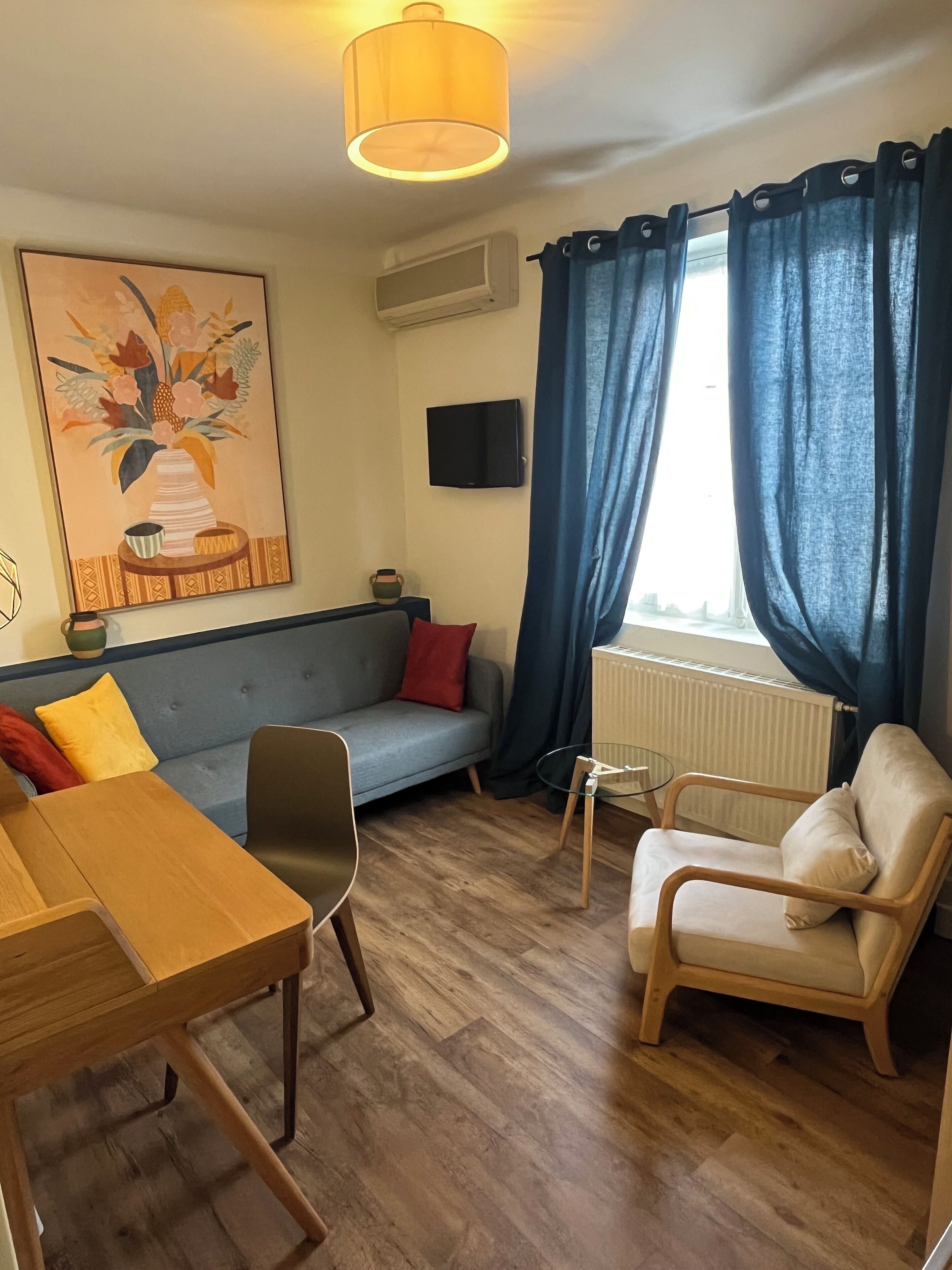 Una sala de estar acogedora con un sofá, una mesa de madera, una mesa baja de vidrio y un sillón color crema. Hay un cuadro en la pared, un televisor montado sobre el sofá y una ventana con cortinas azules.