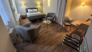 Habitación de hotel con cama, dos sillones, una mesita y un escritorio con silla. Hay lámparas de pared y una ventana con cortinas.
