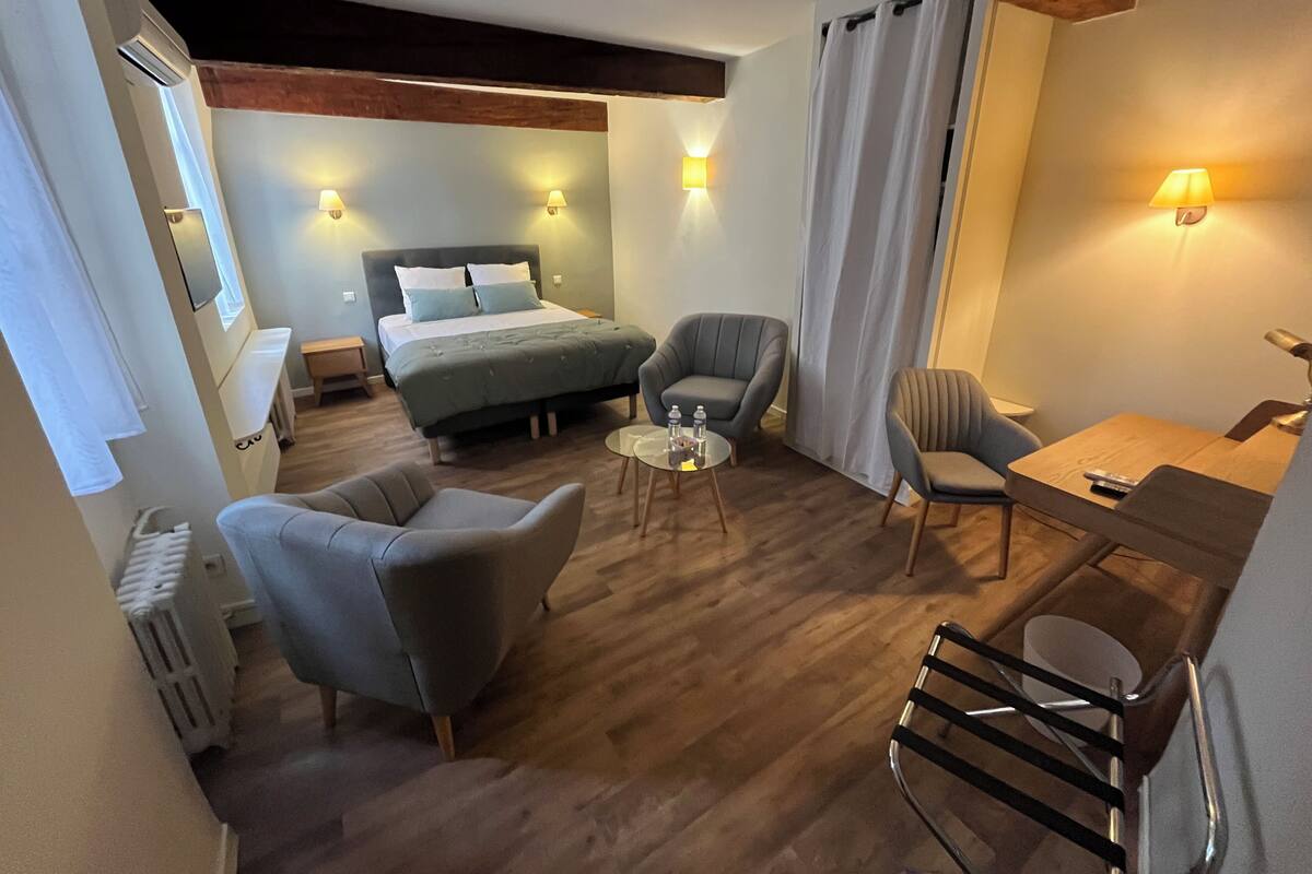 A hotel room with a bed, two armchairs, a small table, and a desk with a chair. There are wall-mounted lamps and a window with curtains.