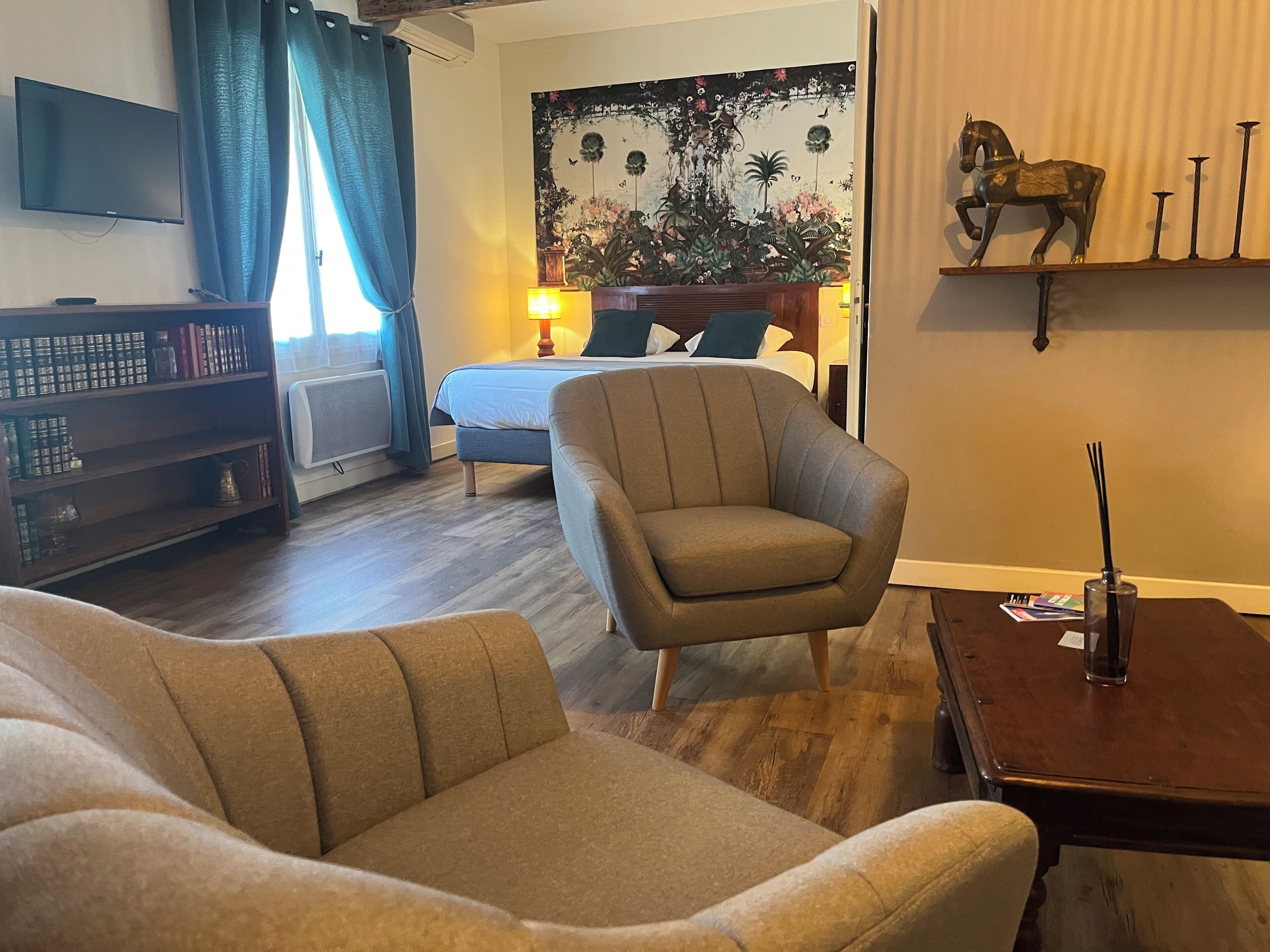 A living room with a sofa, armchair, and a television. There is a bookshelf, a bed, and a decorative wall piece.