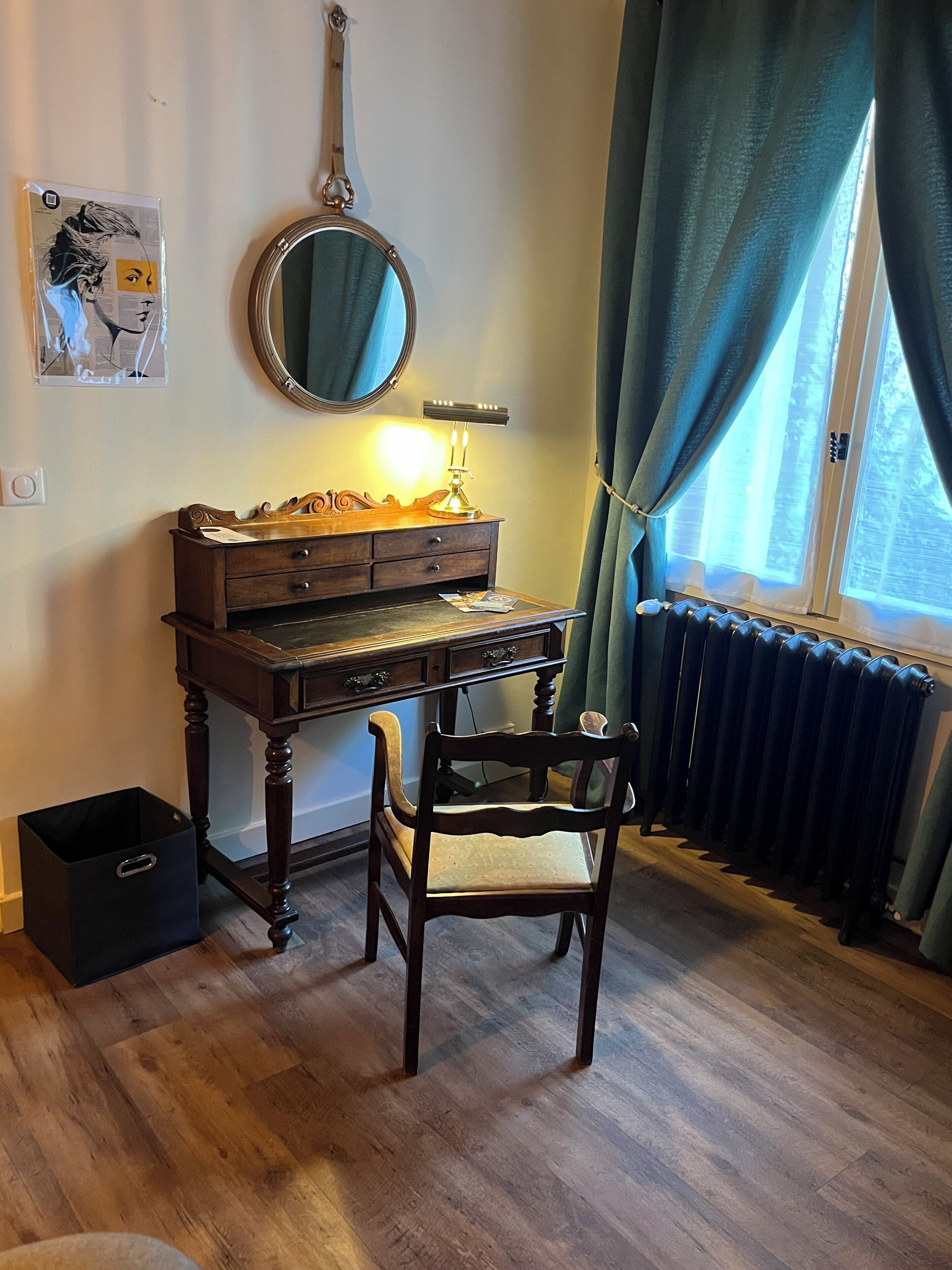 Et træbord med et spejl, en lampe og en stol i et rum med trægulv og et vindue med gardiner.