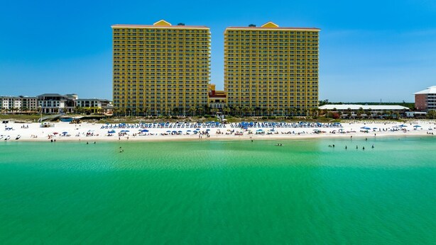 A beachfront hotel with two prominent yellow buildings, a sandy beach with numerous umbrellas, and clear turquoise water.