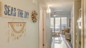 A spacious hallway with a view to a balcony, a 'SEAS THE DAY' sign, and a beaded wall hanging.