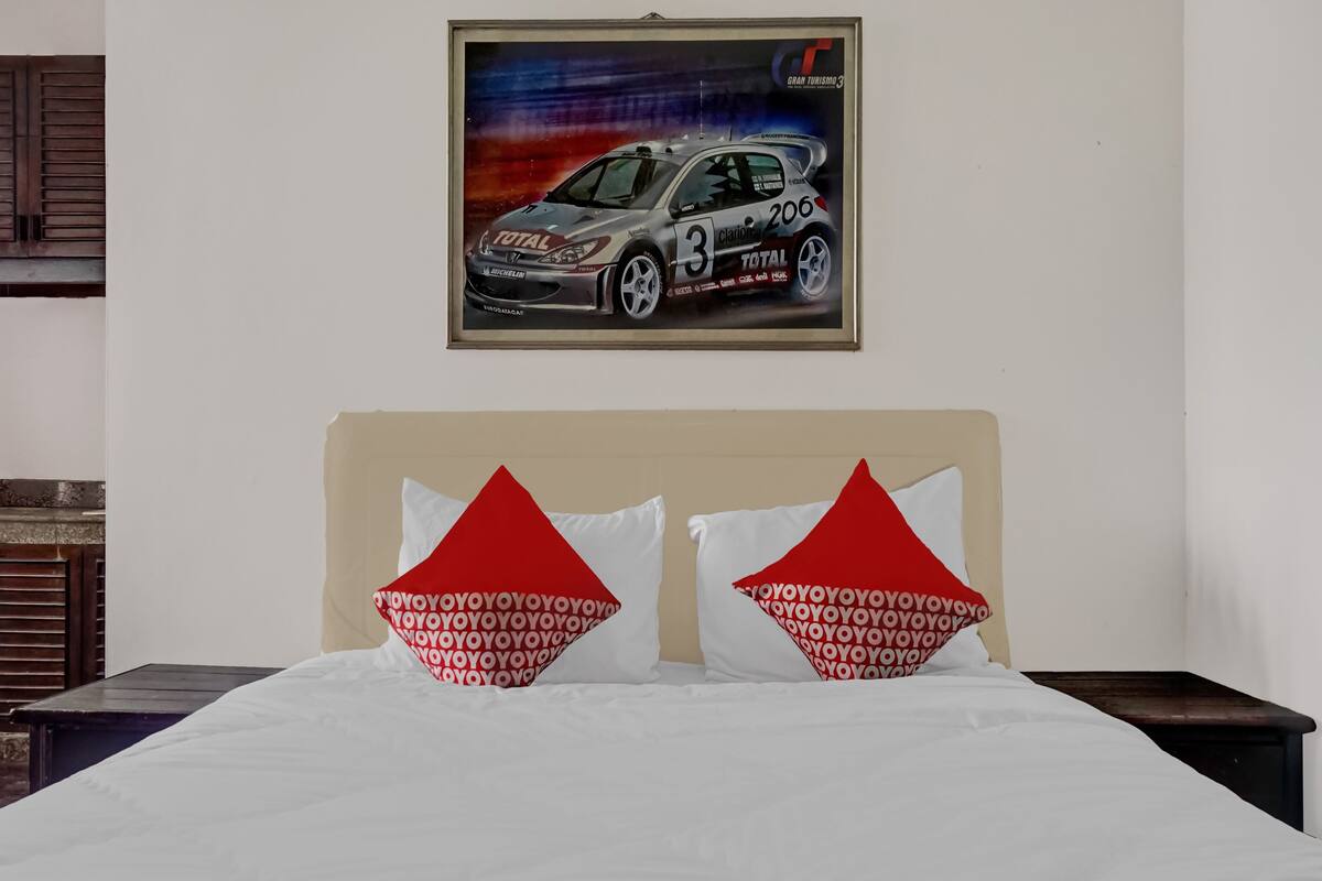 Habitación de hotel con cama, dos almohadas con diseño rojo y blanco, un cuadro enmarcado de un coche en la pared y cabecero de madera.