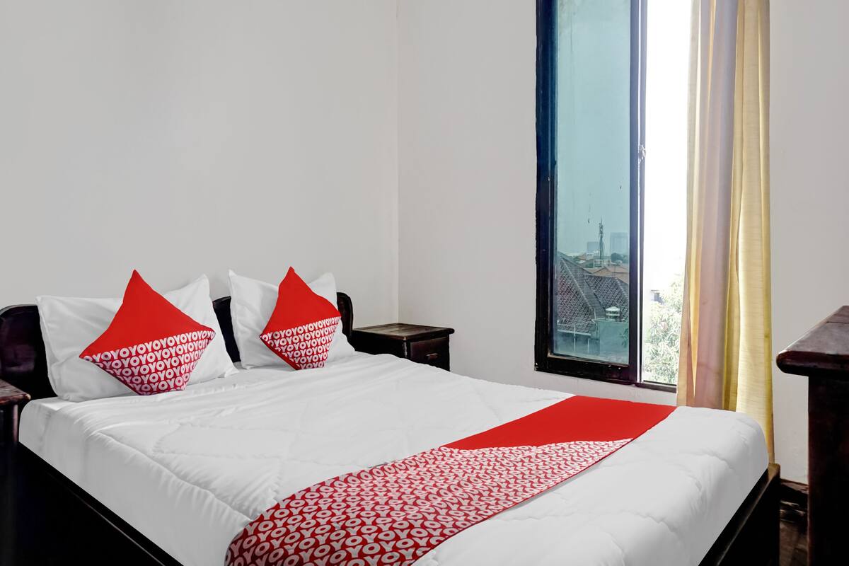 Una cama bien tendida con ropa de cama blanca y roja, vistas de edificios a través de una ventana y una mesita de noche de madera.