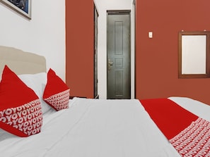 Habitación de hotel con cama, almohadas rojas y blancas, una puerta gris y un cuadro en la pared.