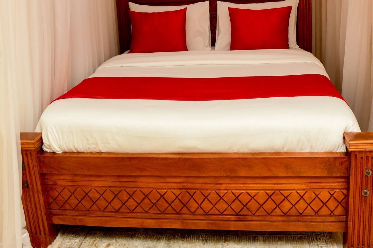 Cama bem arrumada com estrutura de madeira e roupa de cama vermelha e branca, tapete estampado e cortinas leves.