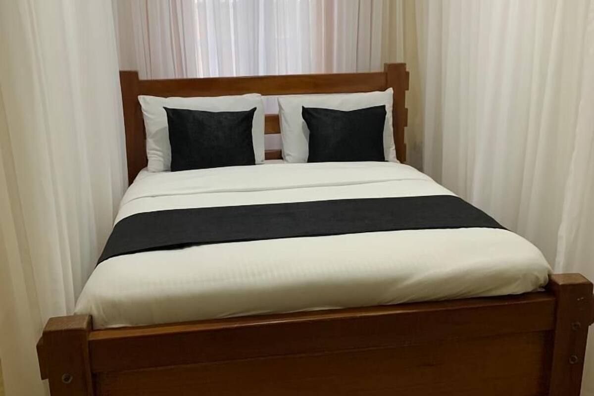 Uma cama de madeira com roupa de cama preta e branca, cabeceira de madeira e um tapete estampado sobre piso de azulejos.