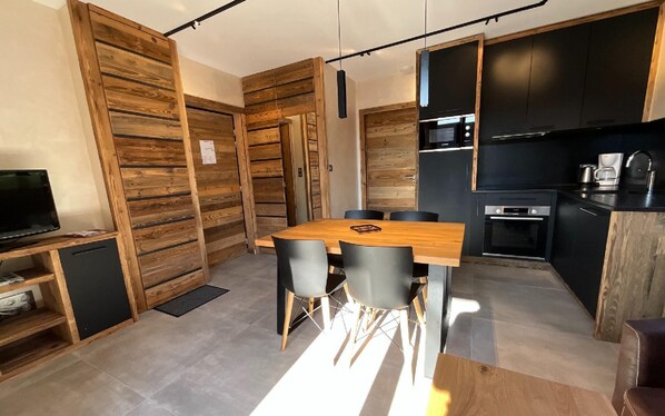 Une cuisine moderne avec des meubles de rangement noirs, une table à manger en bois et un téléviseur posé sur une étagère.