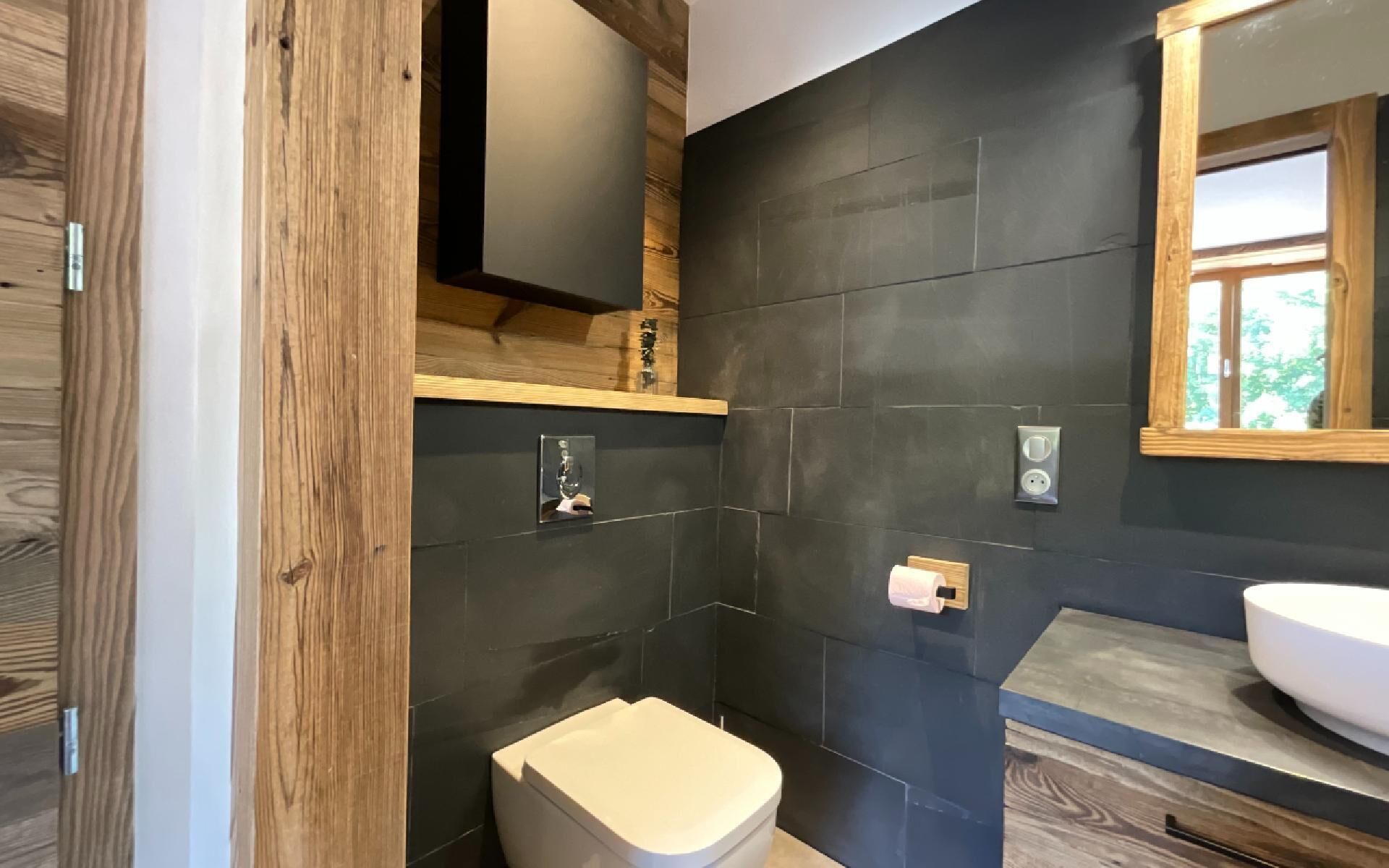 A modern bathroom with dark tiled walls, a wooden vanity, and a toilet.