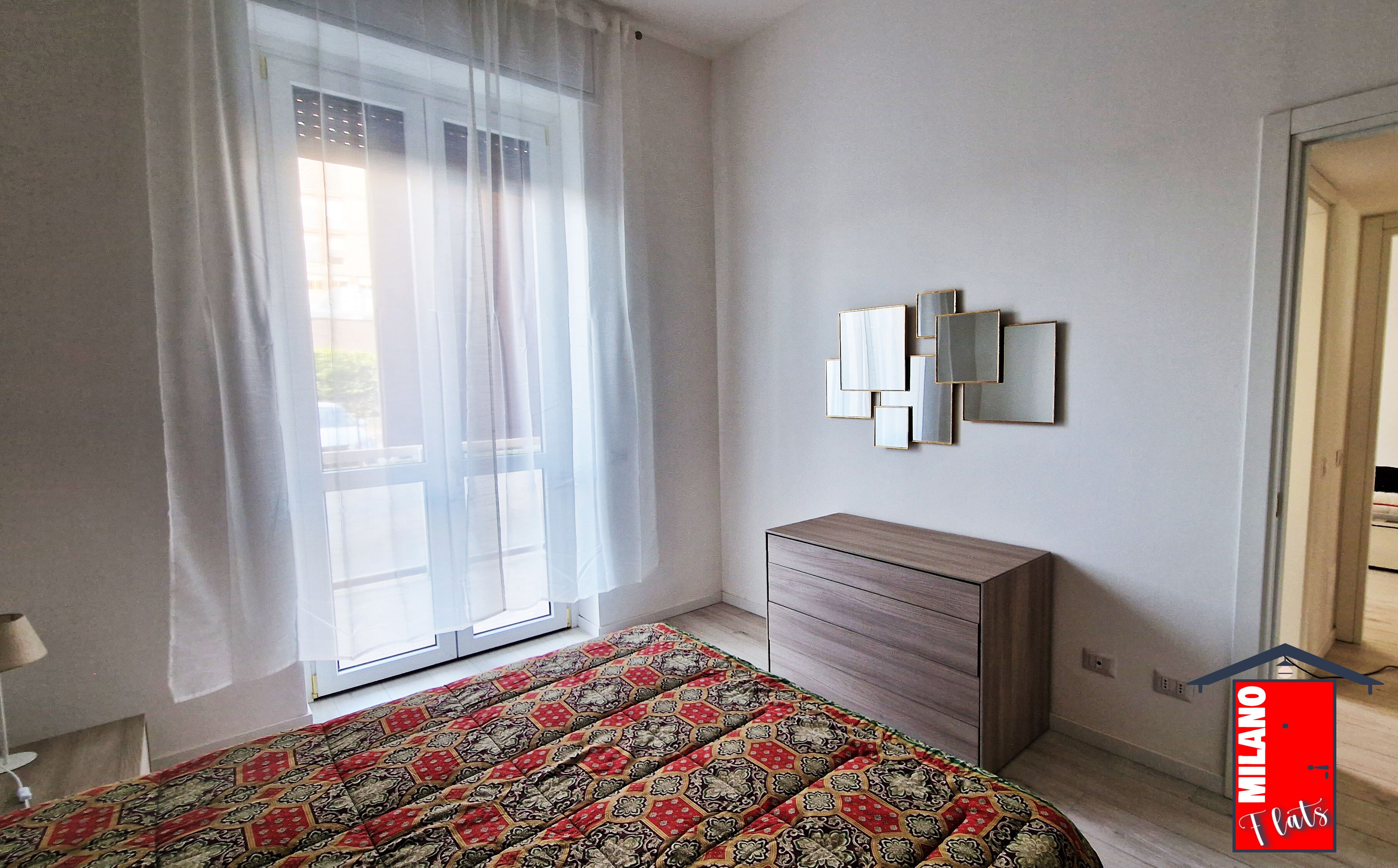 Une chambre à coucher comprenant un lit, une commode, une fenêtre avec des rideaux légers et un miroir fixé au mur.