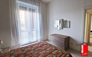 Um quarto com cama, cômoda, janela com cortinas leves e espelho fixado na parede.