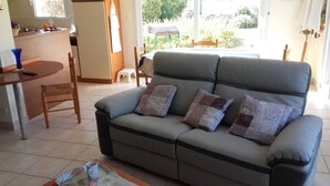 A living room with a sofa, a coffee table, a dining table, and a kitchen area visible through an open door. - Propriété (Saint-Pierre-Quiberon)