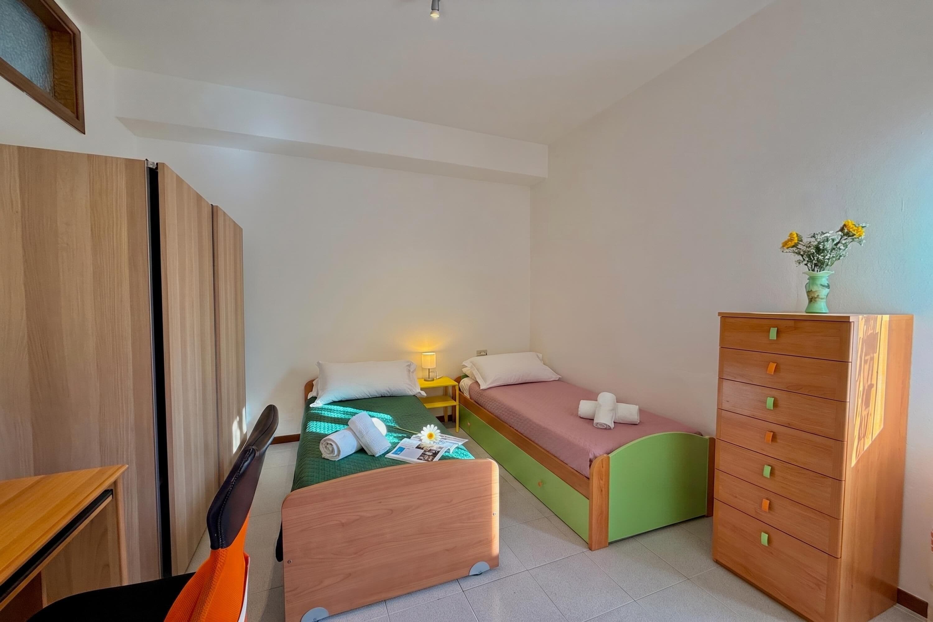 A small, neatly arranged bedroom with a single bed, a desk, a chair, a dresser, and a nightstand.