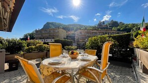 Un área de comedor al aire libre con una mesa preparada para dos personas, rodeada de sillas, y una vista de edificios y colinas al fondo.