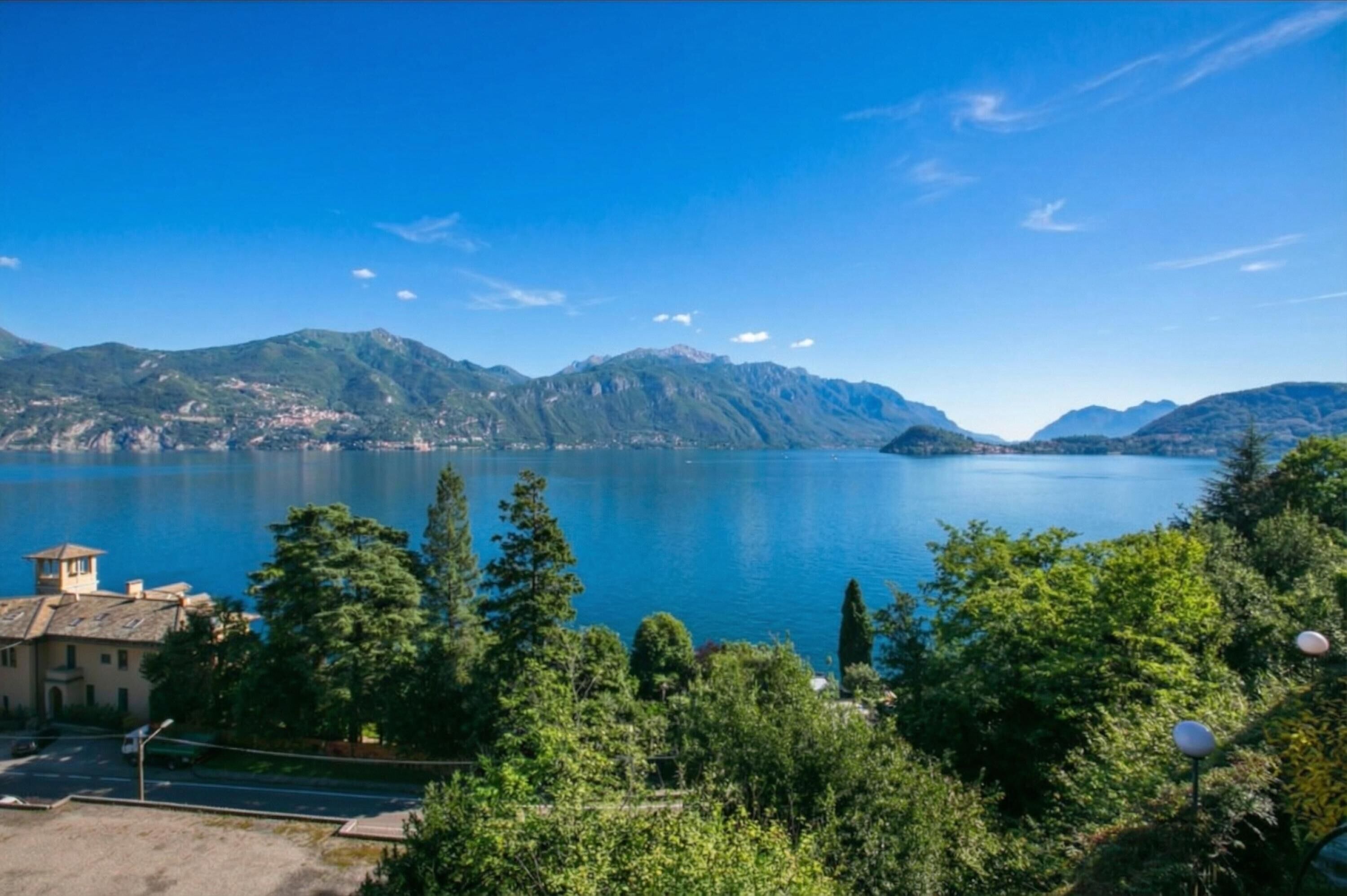 Un lac avec des montagnes en arrière-plan, entouré de verdure et de quelques bâtiments.