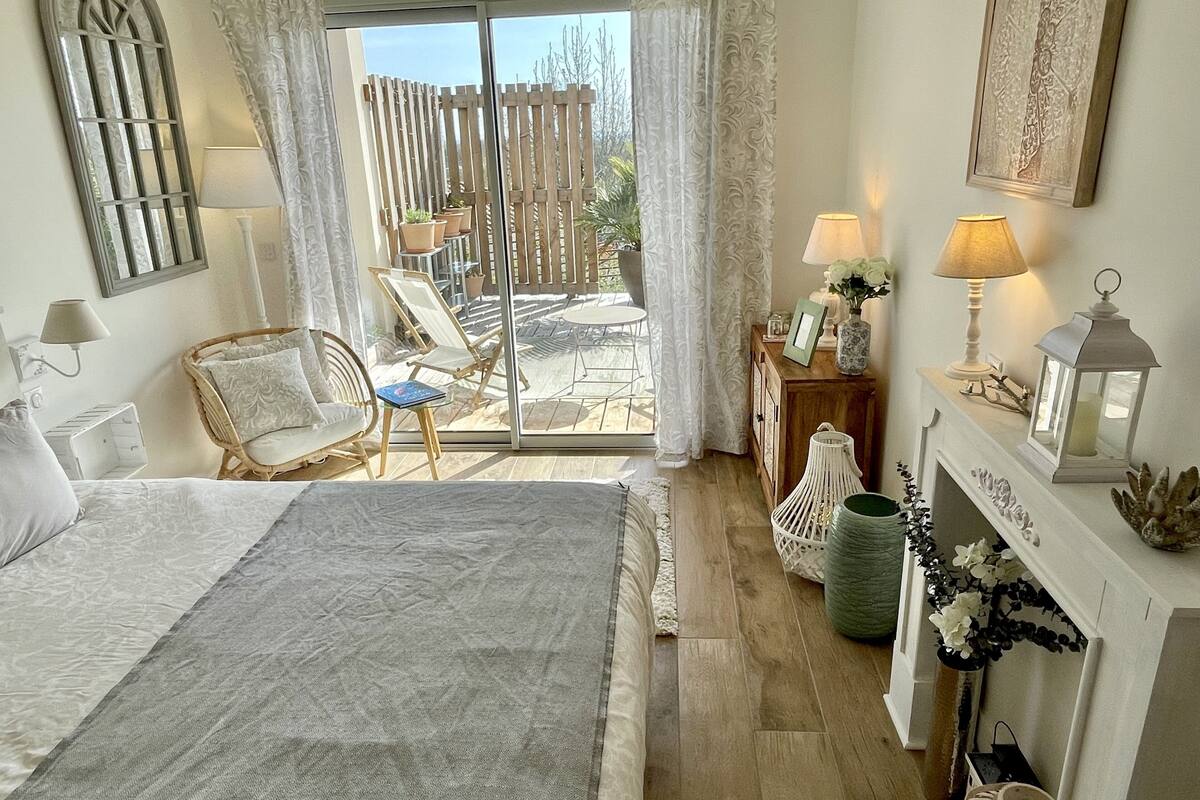 A bedroom with a large bed, a chair, a small table, a fireplace, and a balcony with a view of the outdoors.