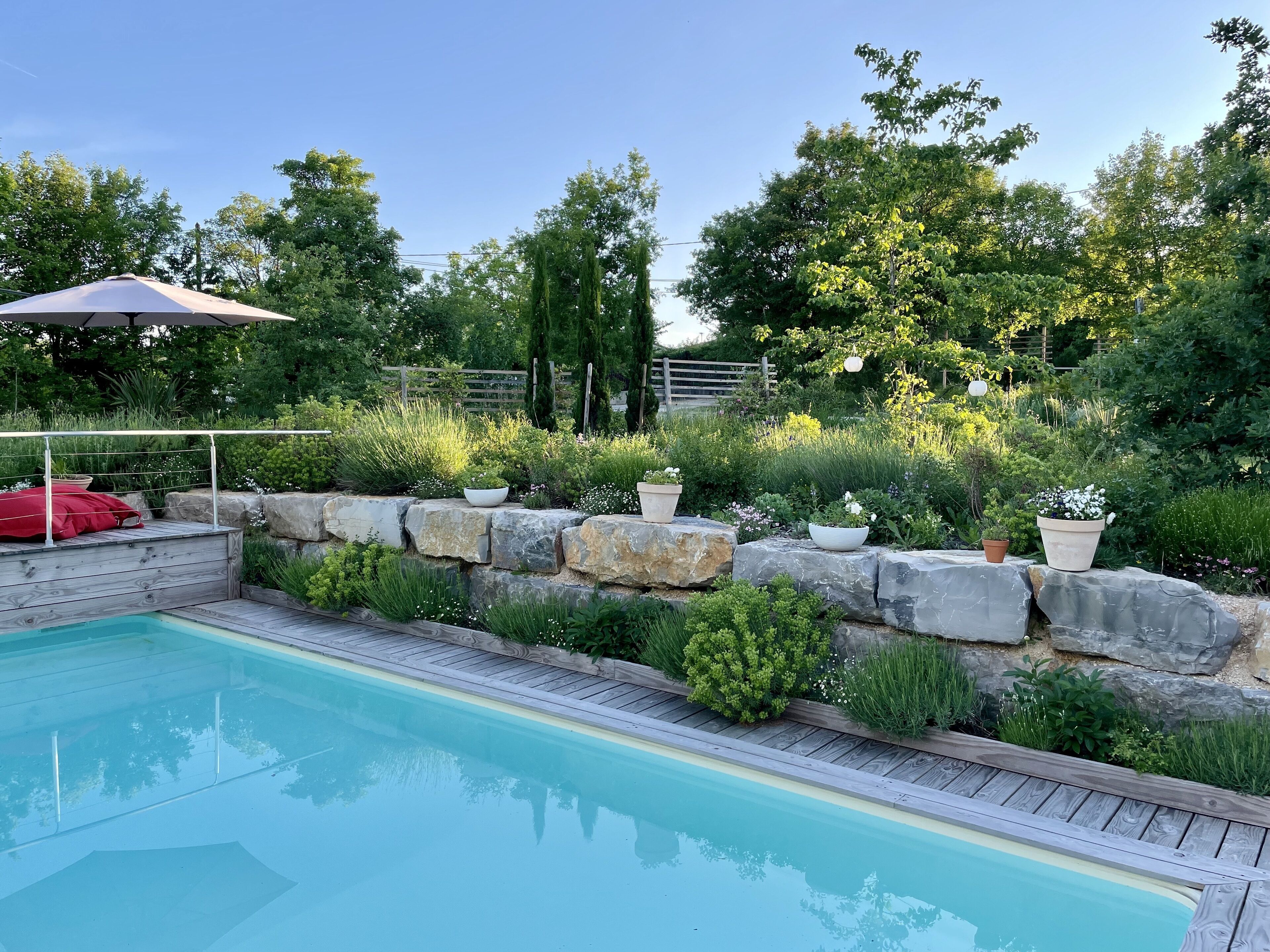 A swimming pool with a wooden deck, surrounded by a stone wall and various plants.