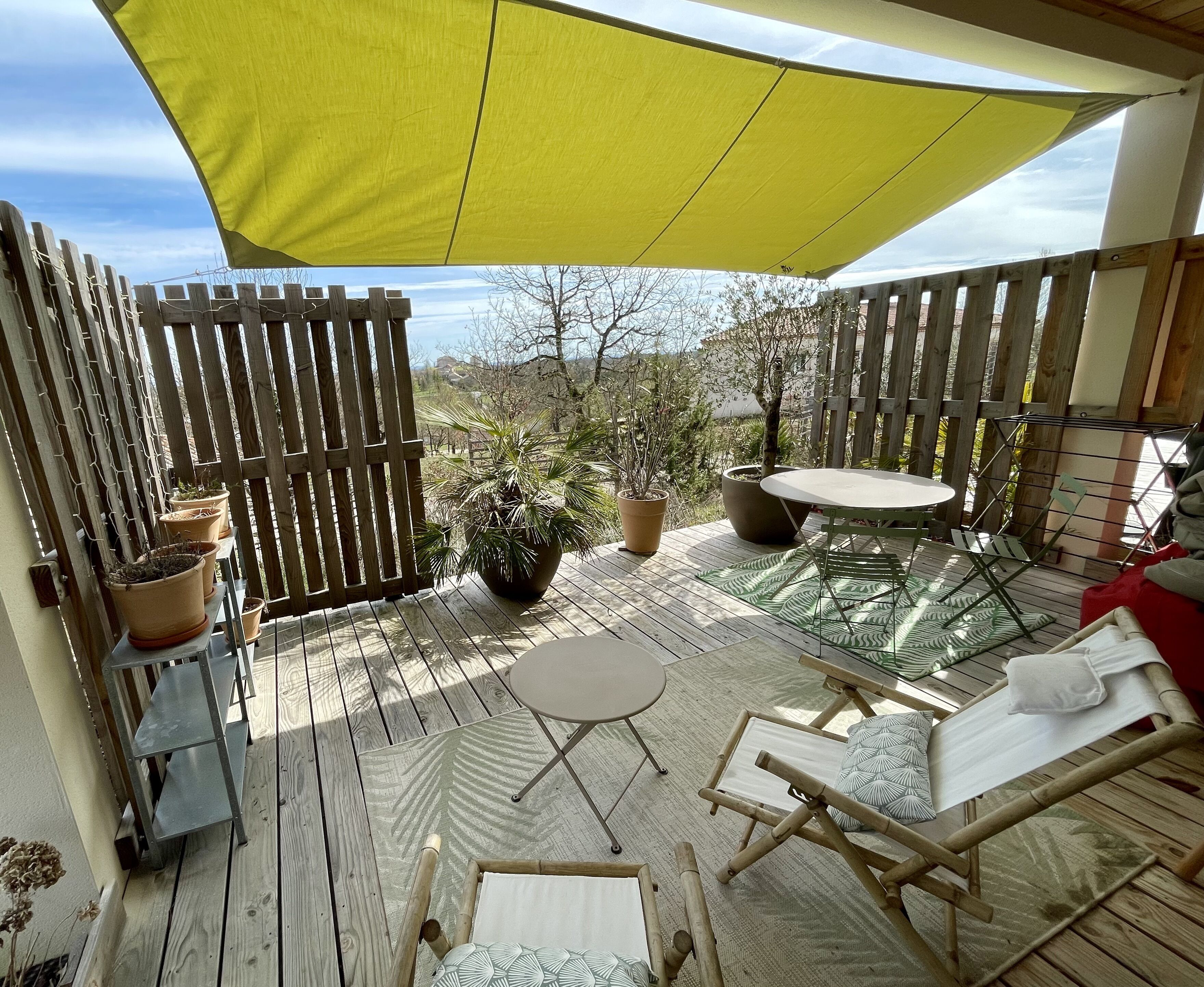 Una terraza de madera con toldo amarillo, plantas en macetas, una mesita y un sillón de relax.