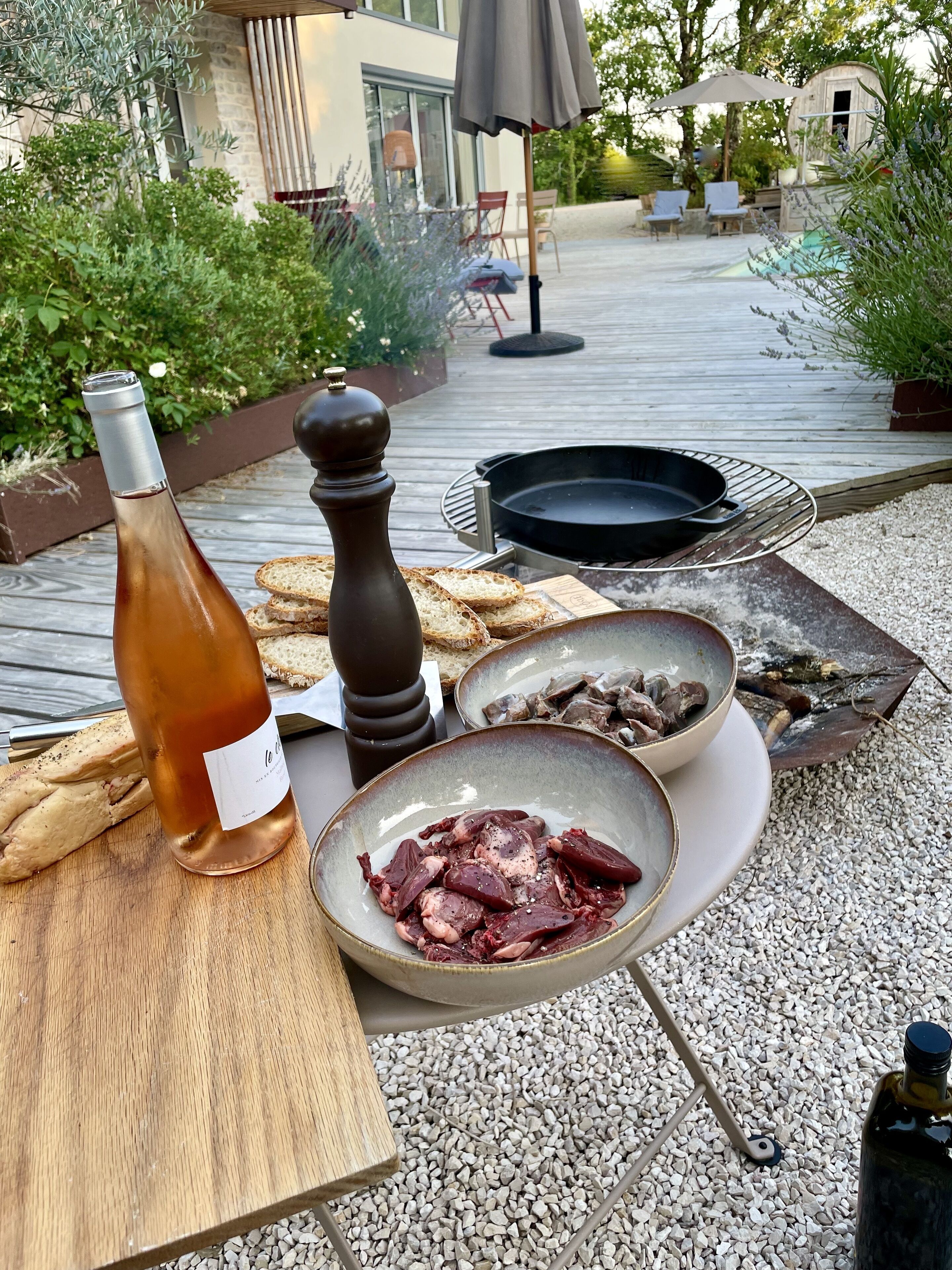 Un espacio de comedor al aire libre con una botella de vino rosado, pan y fuentes de carne sobre una mesa de madera.