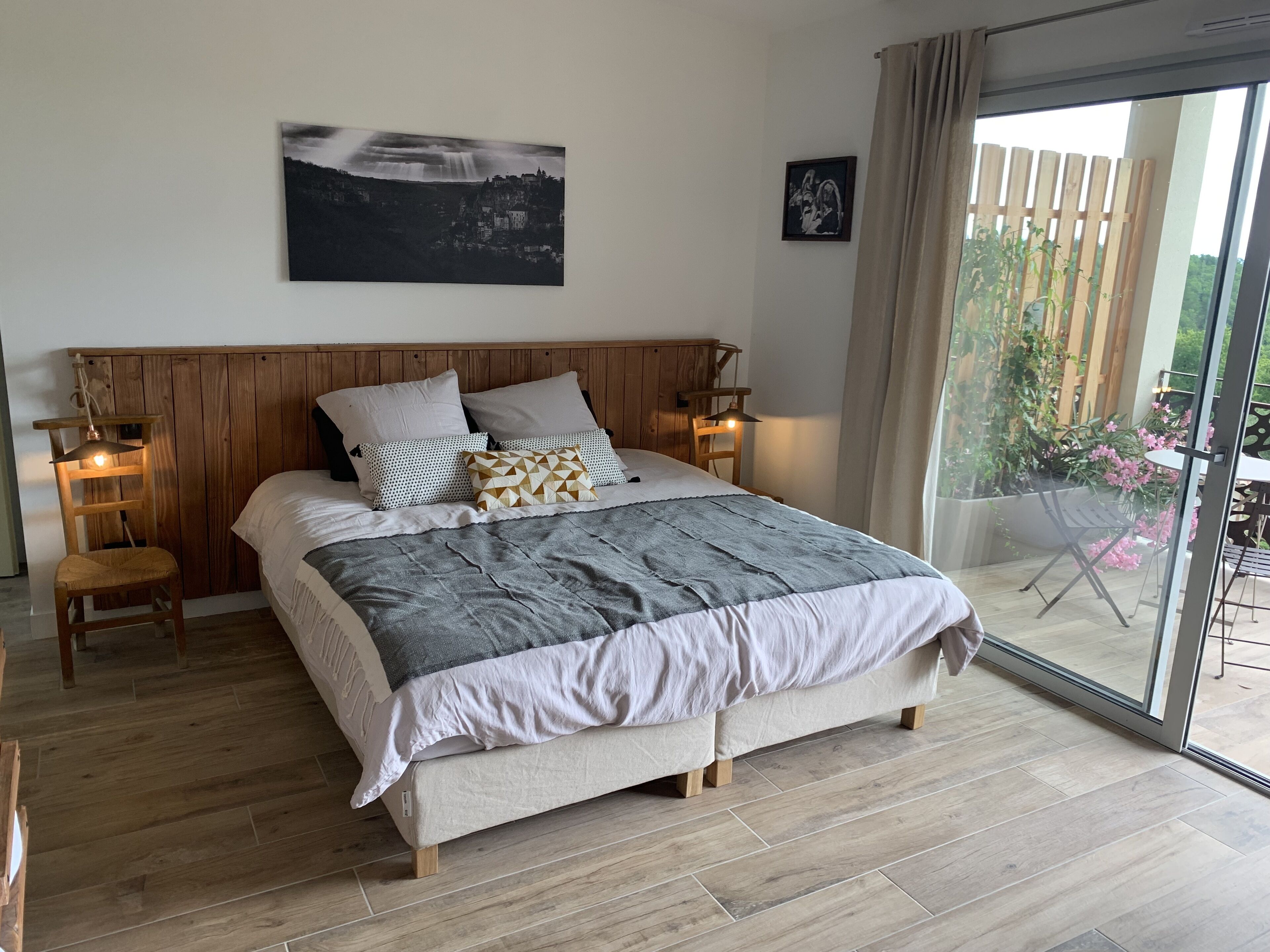 Une chambre à coucher avec une tête de lit en bois, un grand lit recouvert d’une couette grise et blanche, deux tables de chevet avec des lampes, un tableau encadré au mur et une porte-fenêtre coulissante donnant sur un espace extérieur agrémenté de plantes.