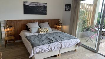 Une chambre à coucher avec une tête de lit en bois, un grand lit recouvert d’une couette grise et blanche, deux tables de chevet avec des lampes, un tableau encadré au mur et une porte-fenêtre coulissante donnant sur un espace extérieur agrémenté de plantes.