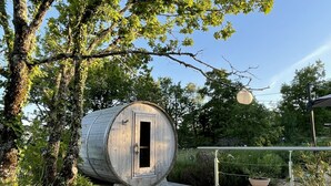Een cilindervormige houten constructie met een deur, omringd door een houten terras en stenen trappen, gelegen in een weelderige tuin met bomen en planten.