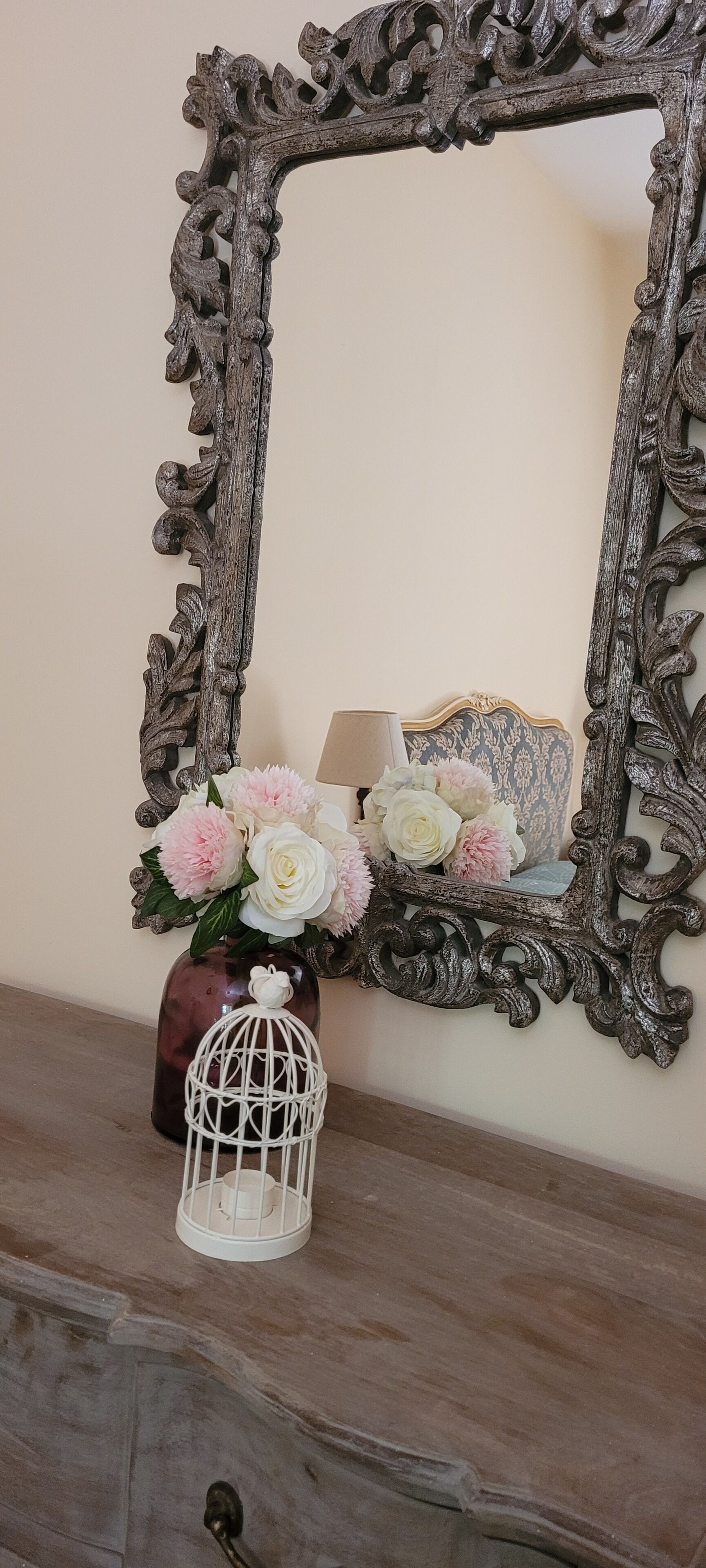 Un espejo con un marco ornamentado, un florero con flores y una jaula de pájaros sobre un aparador de madera.