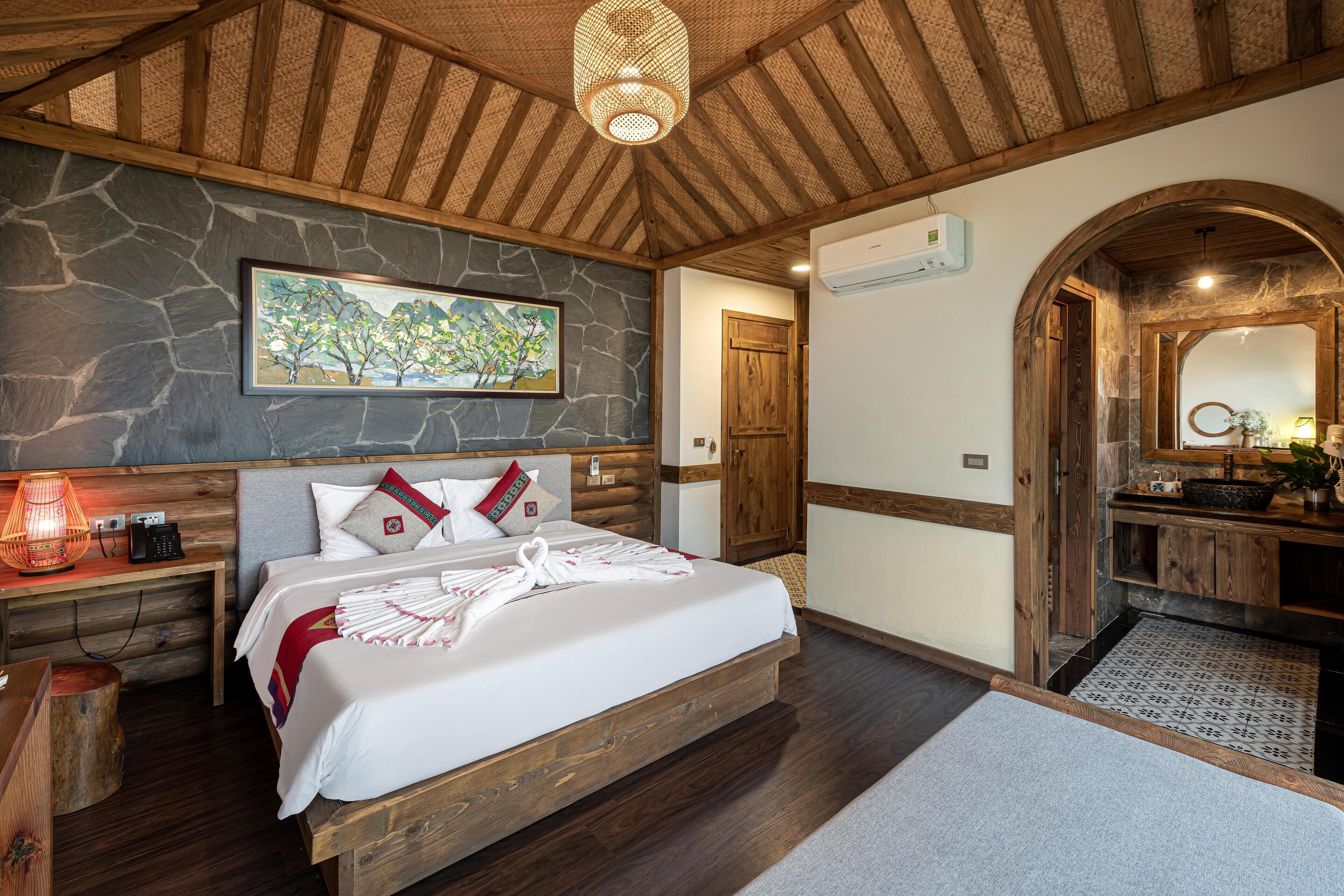 A bedroom with a stone wall, wooden ceiling beams, a bed with white linens, a bedside table with a lamp, and a bathroom with a wooden archway.