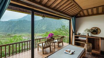 A wooden deck with a view of green hills, a wooden table with a magazine, and a small wooden stool.