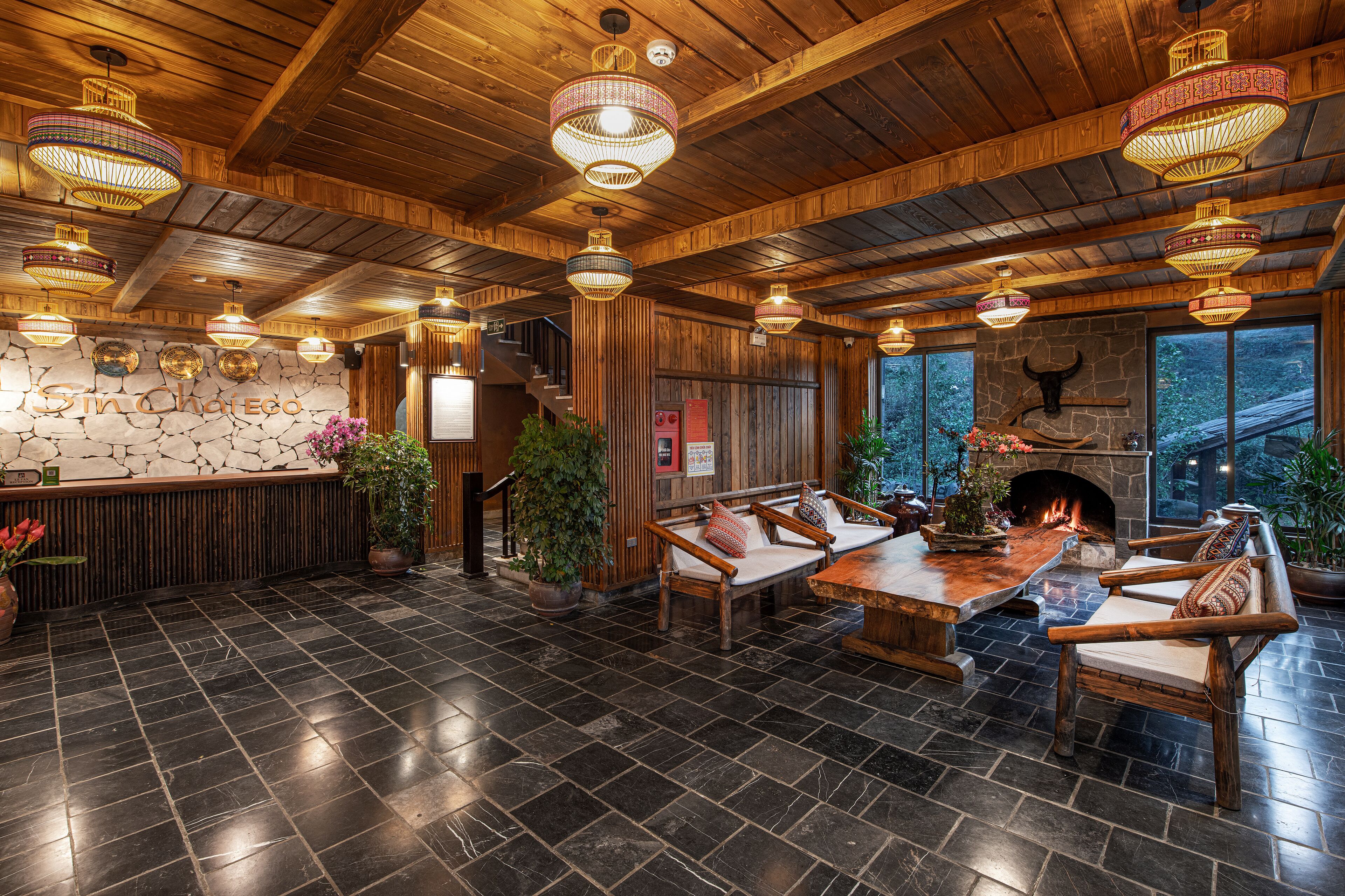 A rustic interior with a stone wall, wooden ceiling, and a fireplace. There are multiple seating areas with wooden furniture and potted plants.