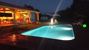 A swimming pool with a wooden deck, a covered patio area, and a house in the background. - Peace and green charm (Salies-du-Salat)