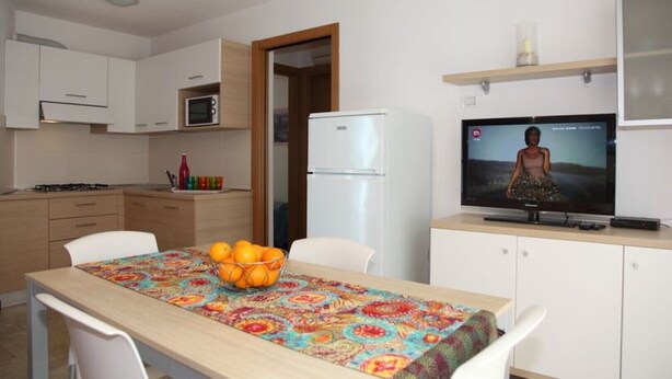 Una cocina moderna con armarios blancos, un microondas, un refrigerador y una mesa de comedor con mantel colorido y un cuenco de naranjas.