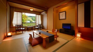 Une pièce de style japonais traditionnel, avec un sol recouvert de tatamis, une petite table basse avec des coussins, un téléviseur et une vue sur un paysage verdoyant, visible à travers une porte coulissante.
