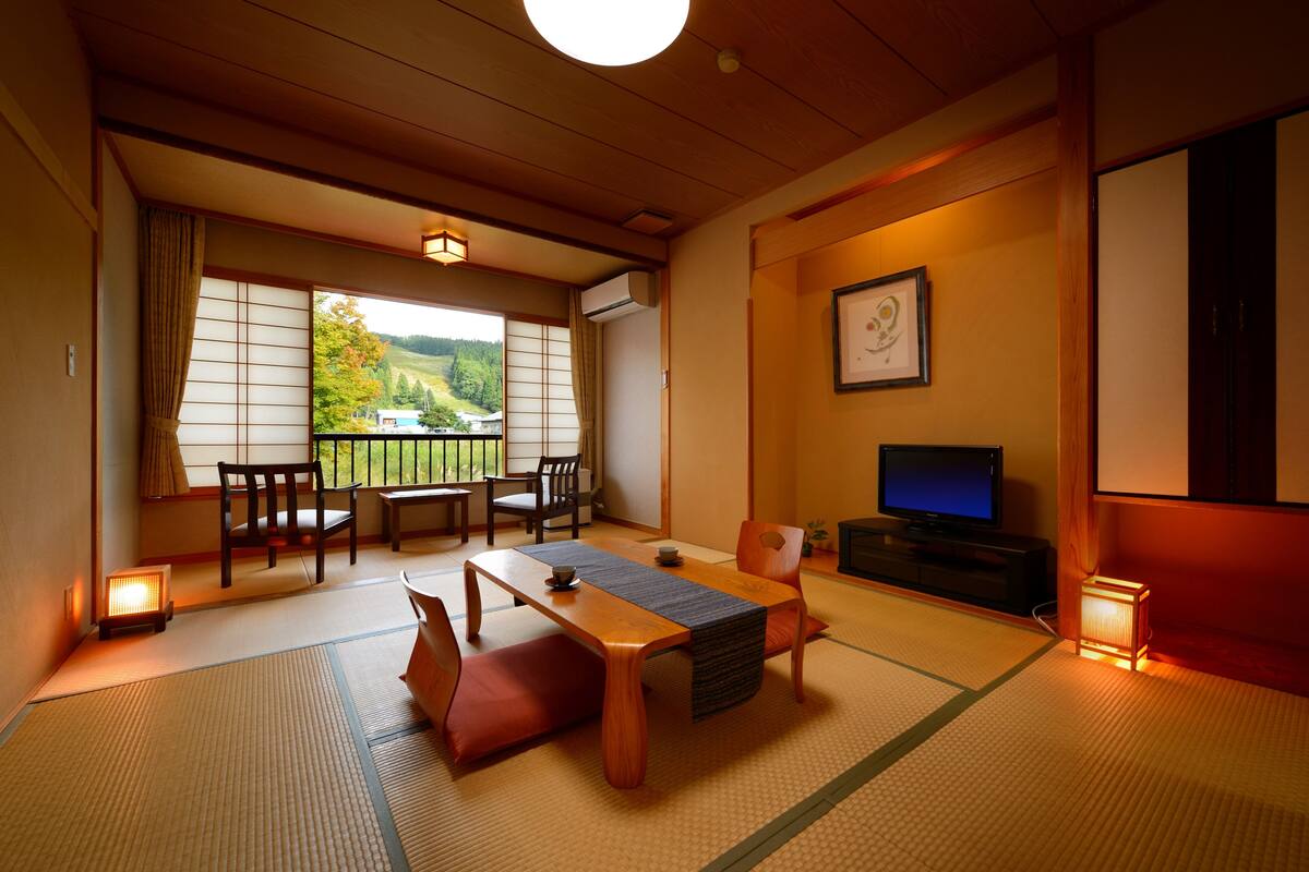 Una habitación japonesa tradicional con suelo de tatami, una mesa baja con cojines, un televisor y vistas a un paisaje verde a través de una puerta corredera.