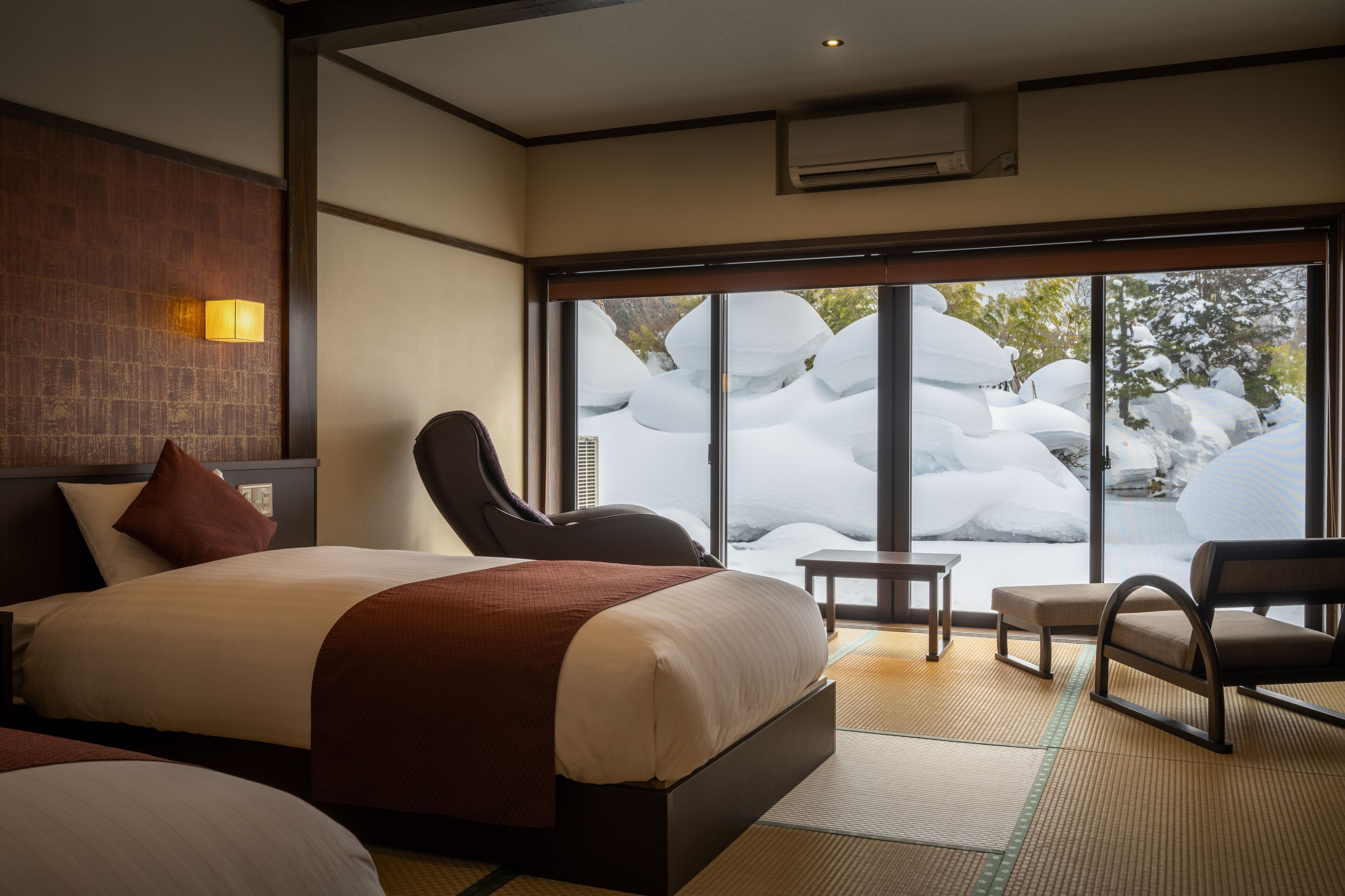 A hotel room with a large bed, a chair, a small table, and a view of snow-covered rocks through large windows.