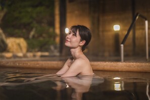 Une personne est assise au bord d’une piscine, dont le rebord est en bois, avec une rambarde métallique en arrière-plan.
