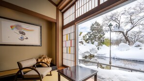 Une pièce de style japonais traditionnel, avec un sol recouvert de tatamis, une table basse et une chaise. Une grande fenêtre donne sur un paysage enneigé, avec un arbre et un lampadaire.