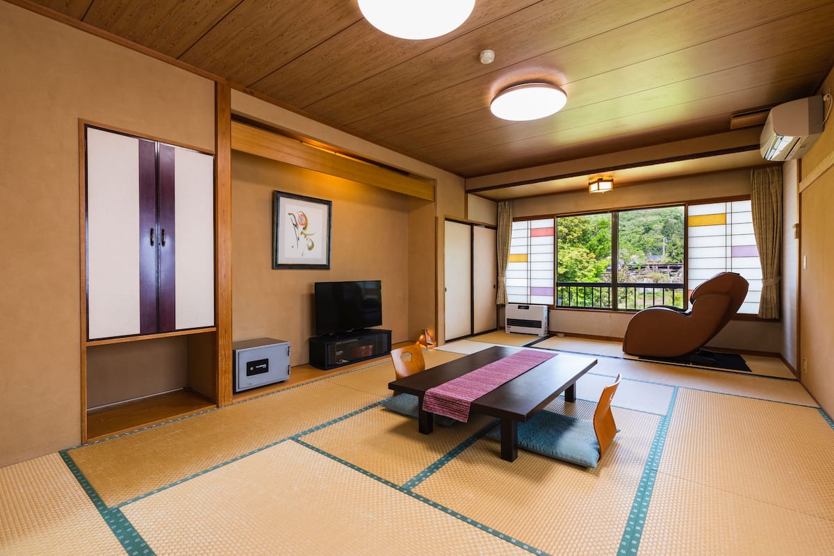 Una habitación japonesa tradicional con suelo de tatami, una mesa baja con un tapete rosa, un televisor y una puerta corredera que da a un balcón con vistas a la vegetación.