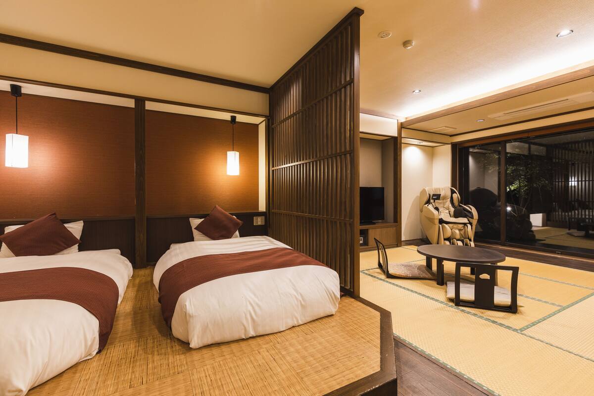 Habitación de hotel con dos camas, una zona de estar con mesa y sillas, y vistas al exterior a través de amplios ventanales.