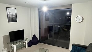 Een moderne woonkamer met een flatscreen televisie op een glazen tafel, een zwarte bank met een blauw kussen en een glazen salontafel.