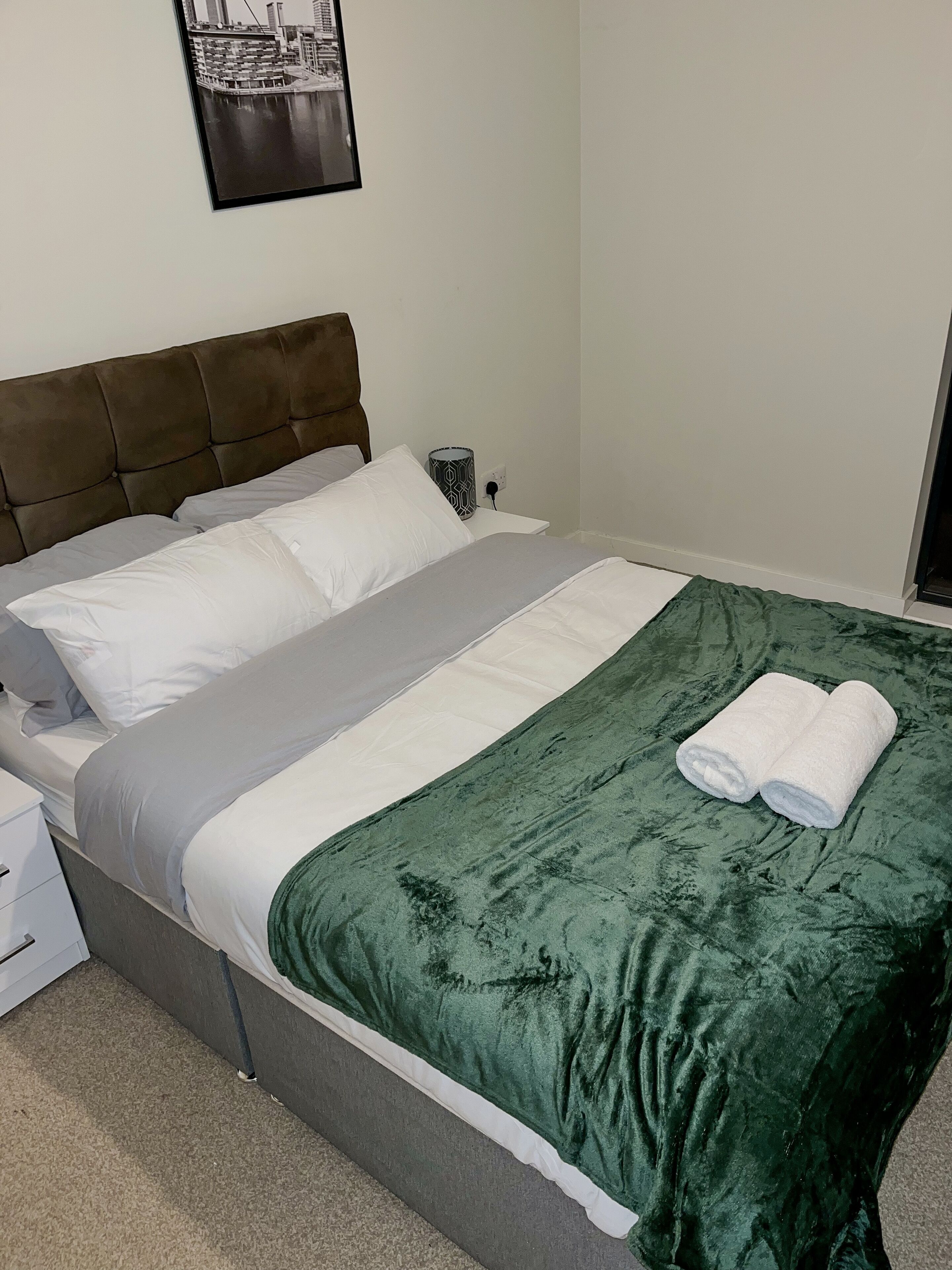 Uma cama bem arrumada com um cobertor verde e almofadas brancas, uma mesa de cabeceira com abajur e um quadro na parede.