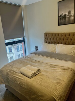 A hotel room with a large bed, a window with a view of buildings, a headboard, and a framed picture on the wall.