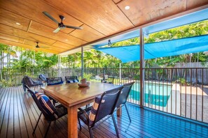 Eine Terrasse im Freien mit Holztisch und -stühlen, einem Deckenventilator und Blick auf einen Swimmingpool.