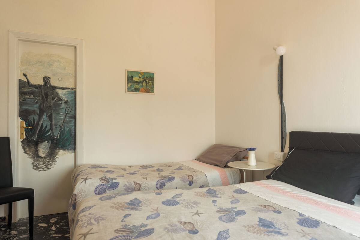 A room with two beds, a small table with a lamp, and a painting on the wall.